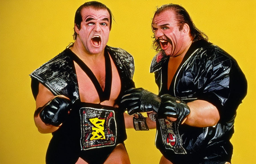 Professional wrestlers with championship belts in black outfits and face paint on yellow backdrop