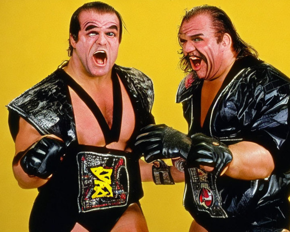 Professional wrestlers with championship belts in black outfits and face paint on yellow backdrop