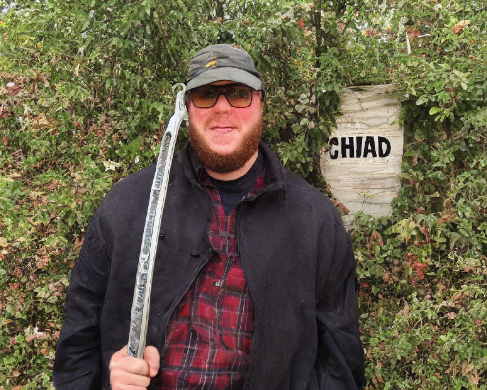 Bearded man in sunglasses and cap with "CHIAD" sign in front of dense foliage