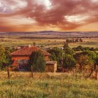 Tranquil dusk landscape with warm sunlight, house, fields, hills, chapel, dramatic sky