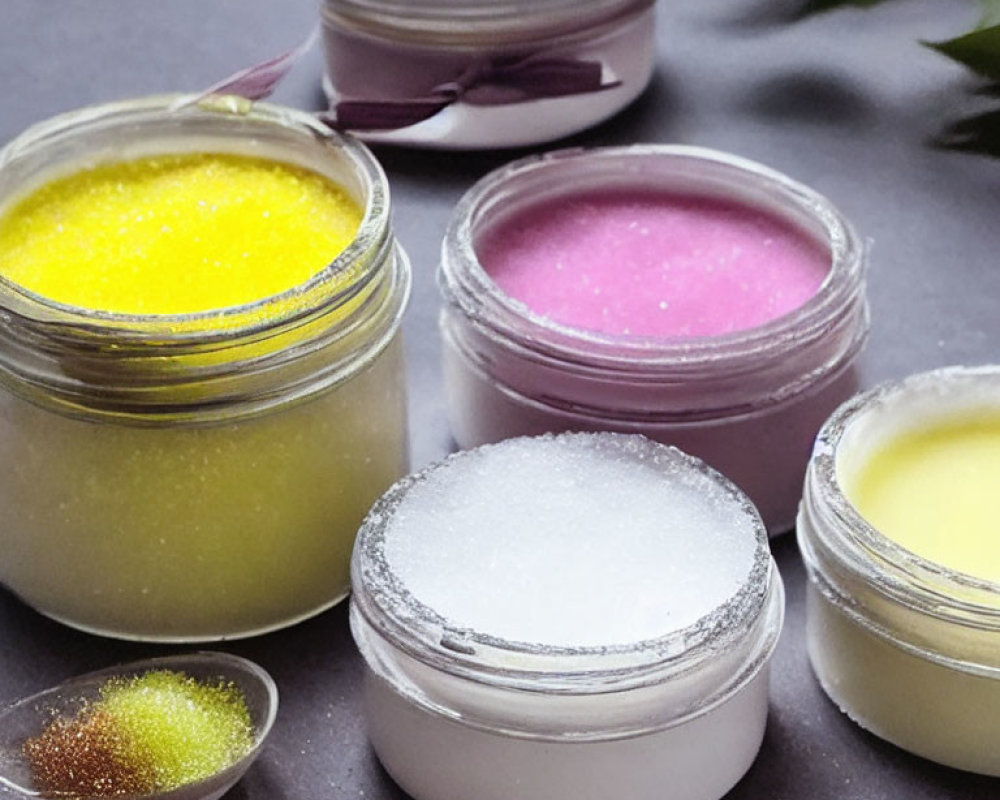 Colorful Lip Scrubs in Open Jars on Gray Surface with Granules and Green Leaves