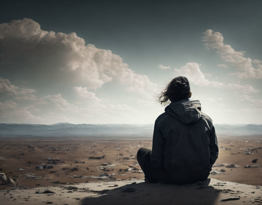 Person sitting on ground gazes at vast desert landscape under cloudy sky