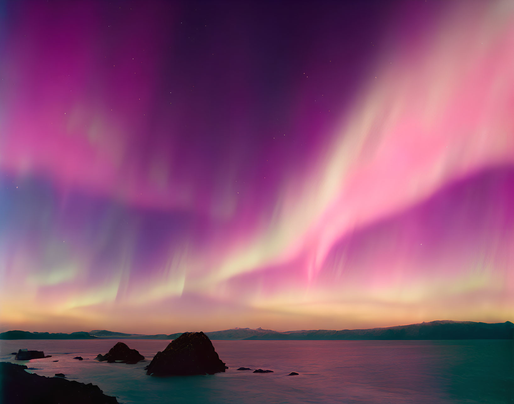 Colorful Northern Lights dance over calm ocean with silhouetted rocks & distant mountains.