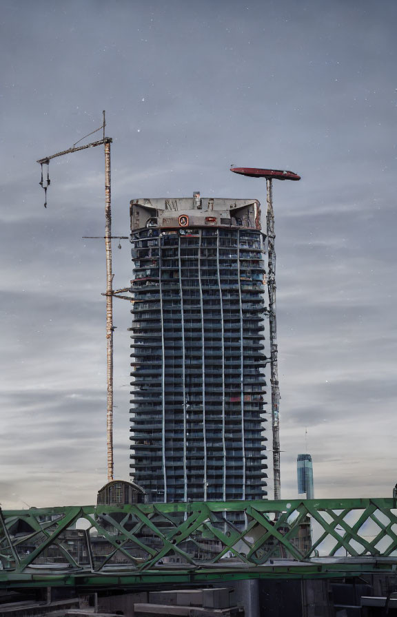 Tall skyscraper under construction with crane at twilight viewed from green bridge