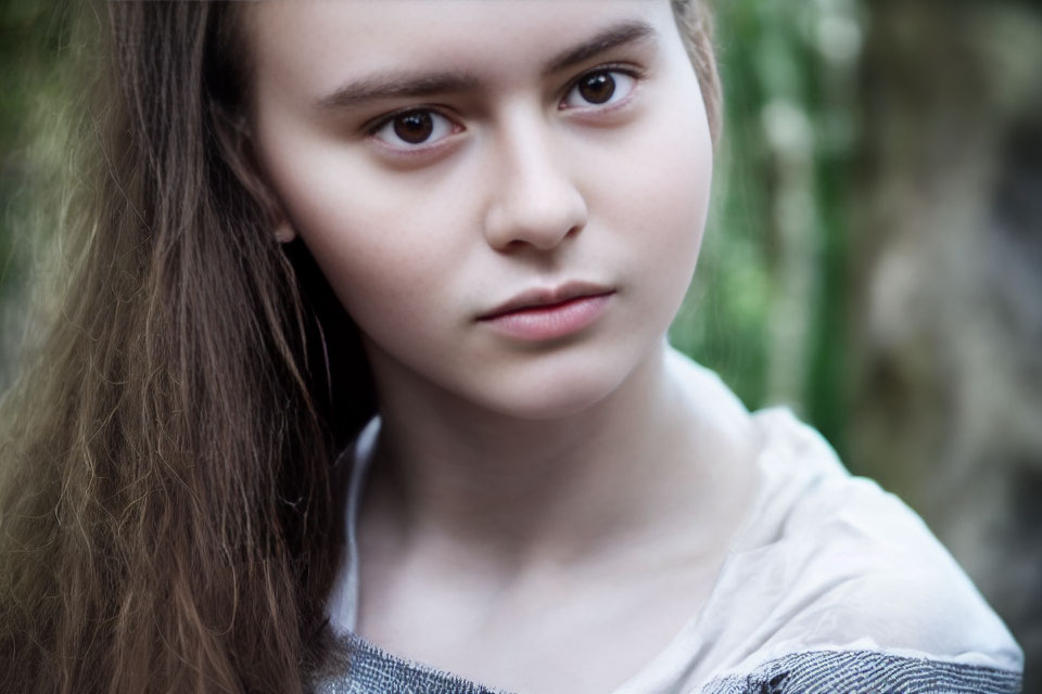 Young woman with long hair in light blouse, thoughtful expression, against blurred natural background