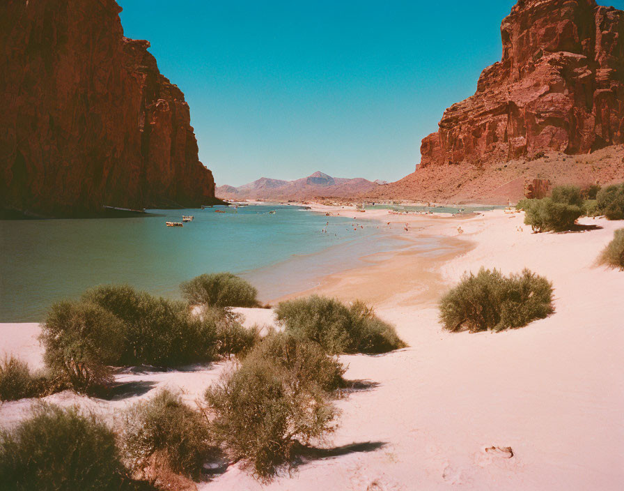 Desert river landscape with sandy shore, people, red rocky cliffs, and clear blue sky