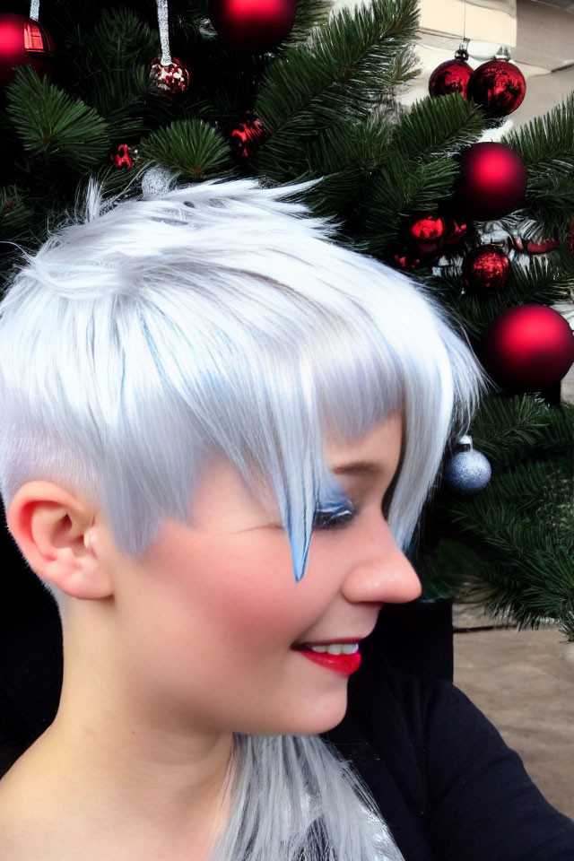 Person with short white hair smiles in front of red and silver Christmas tree