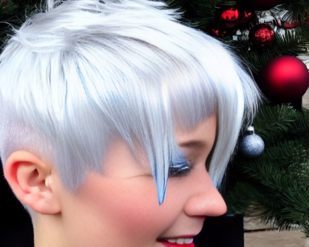 Person with short white hair smiles in front of red and silver Christmas tree