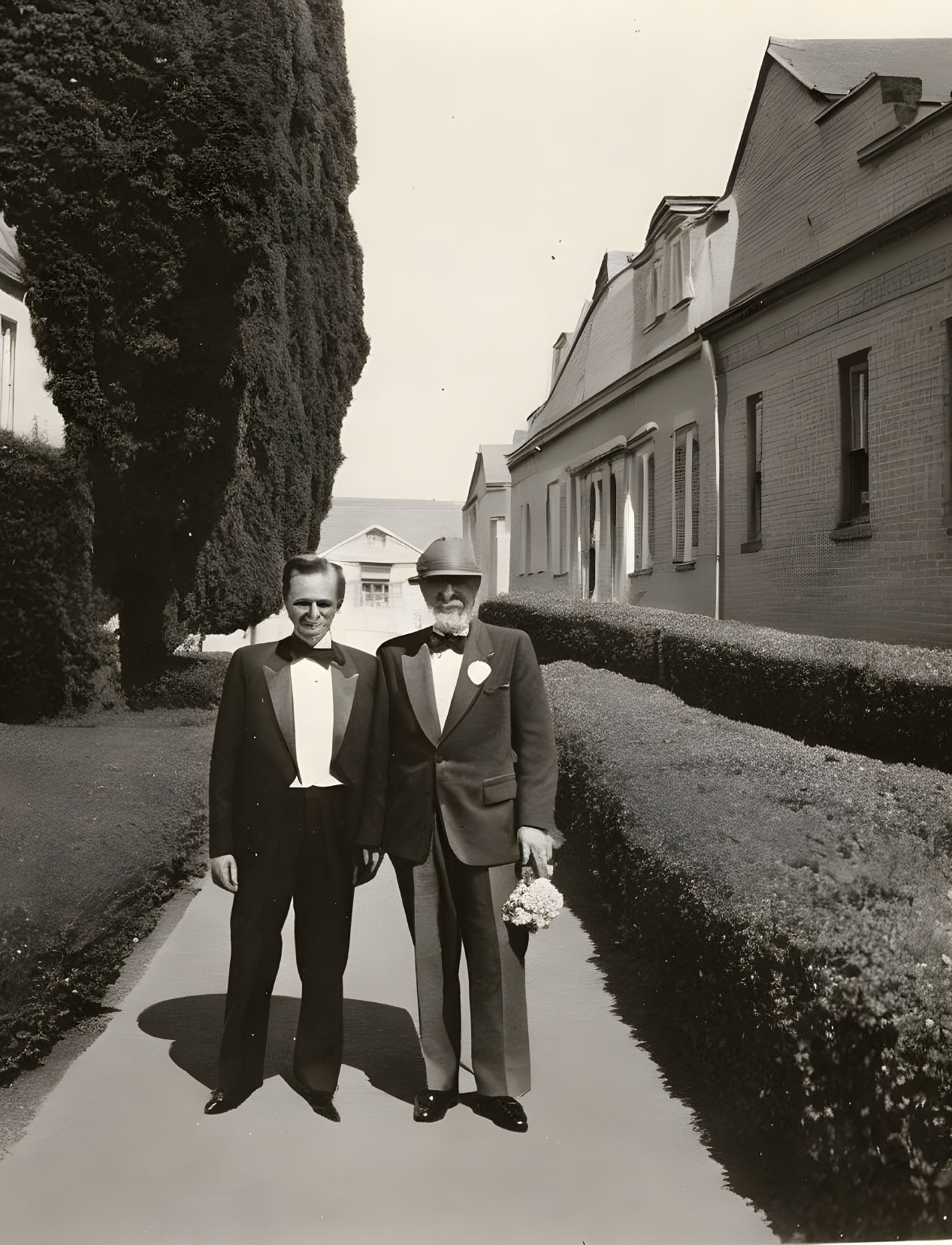 Men in formal attire on pathway with trimmed hedges & buildings.