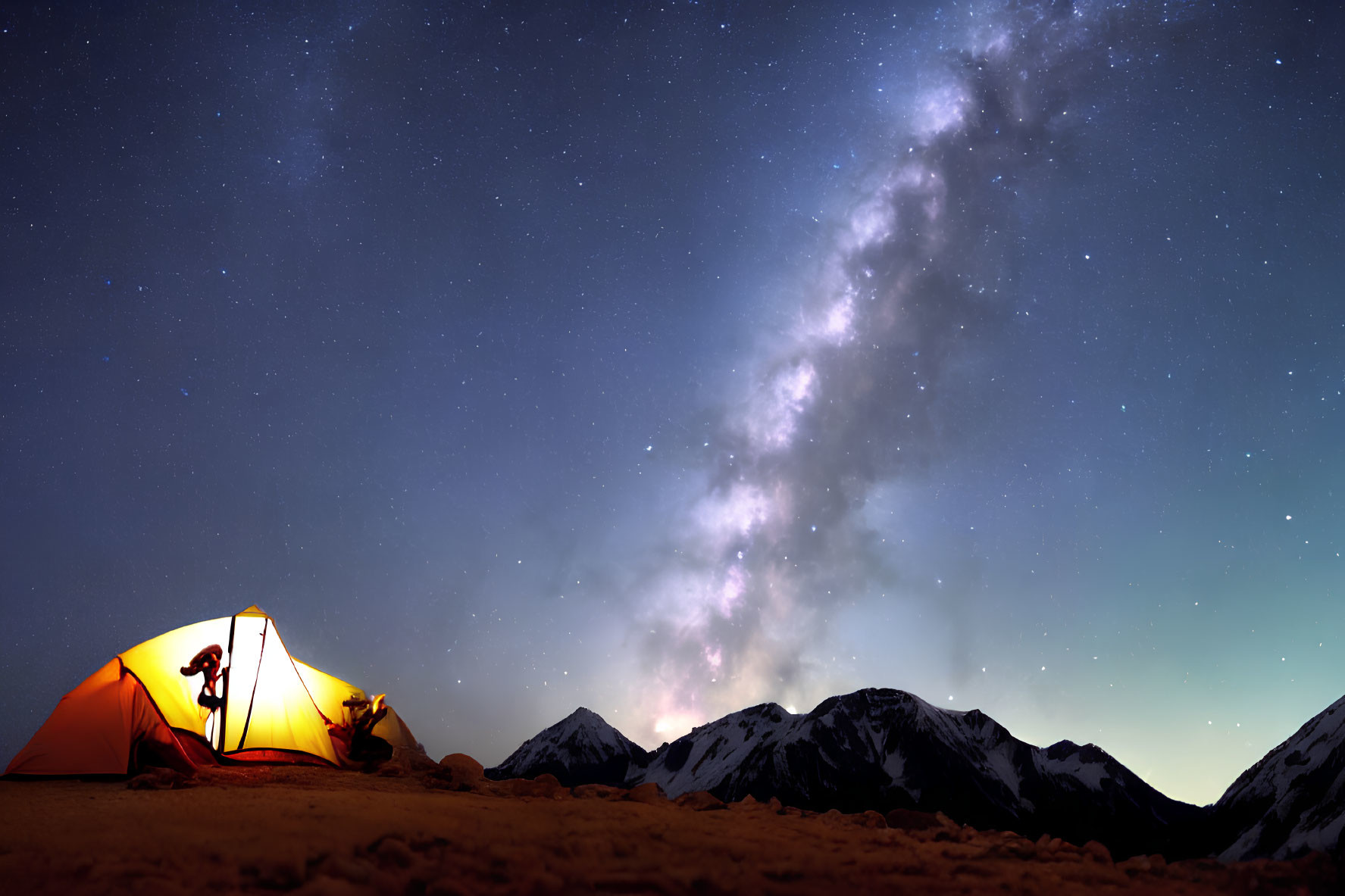 Silhouetted person under glowing tent with starry sky and Milky Way over dark mountains.