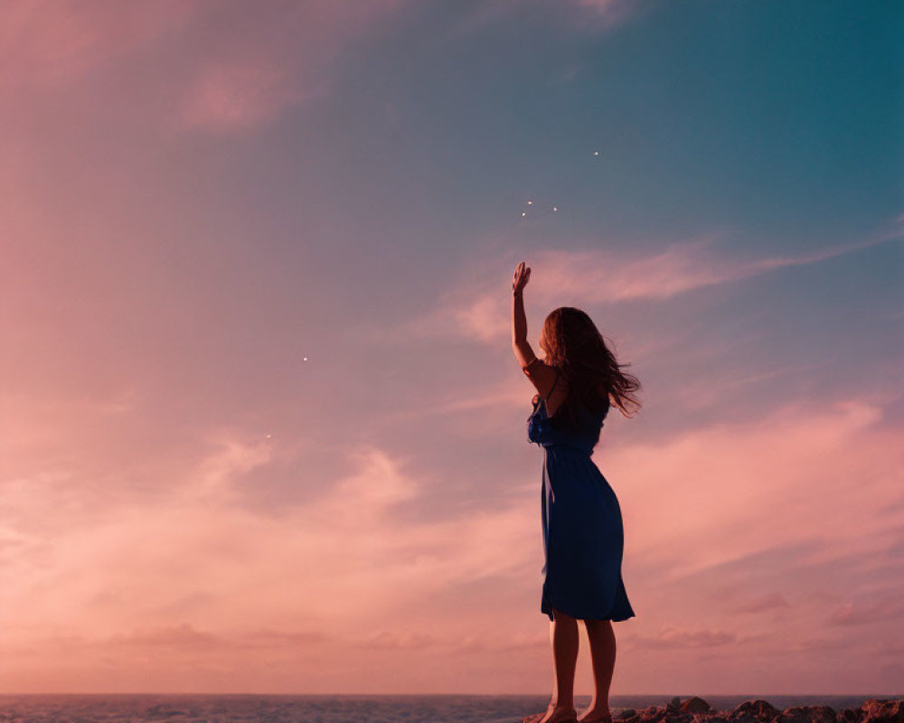 Person in Blue Dress Reaching on Rocky Ocean Terrain at Dusk