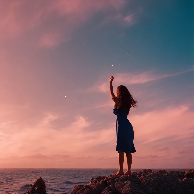 Person in Blue Dress Reaching on Rocky Ocean Terrain at Dusk