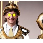 Person in ornate gold and white costume with steampunk-style helmet and glowing orange lenses next to