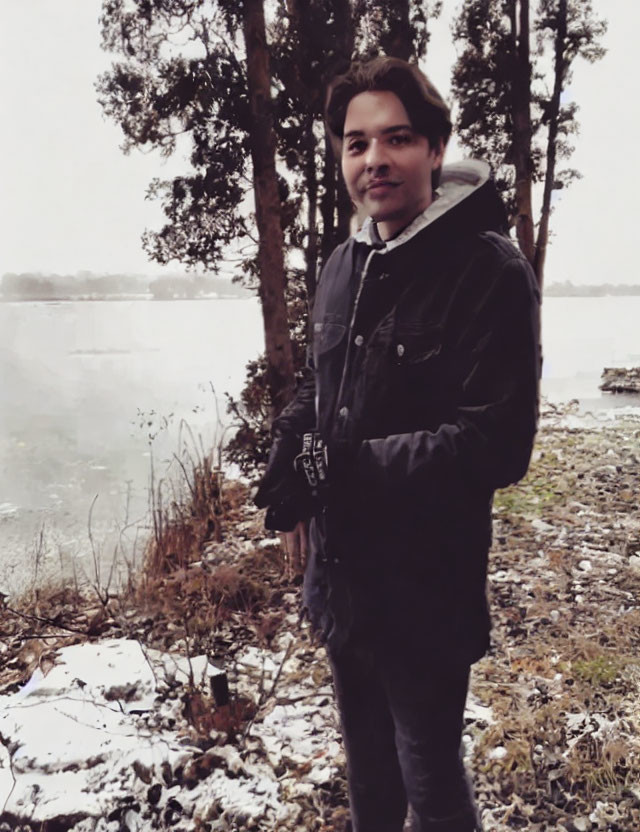 Person in Winter Jacket by Snowy Lakeside with Trees and Overcast Skies