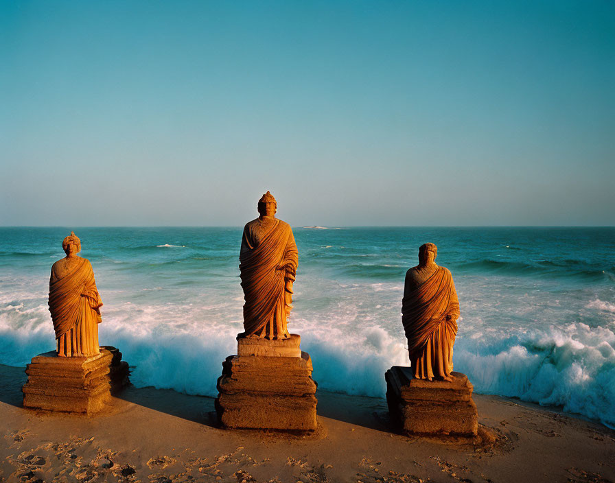 Statues of robed figures by the sea with crashing waves under clear sky