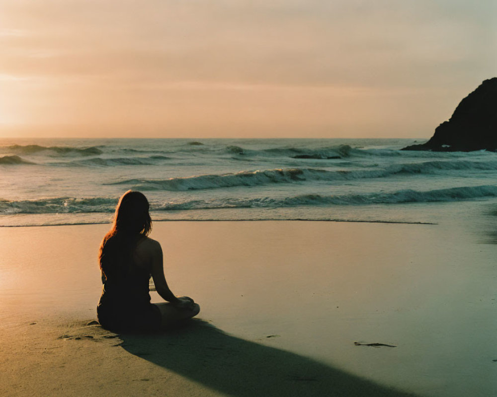 Person sitting on sandy beach at sunset with shadow stretching towards ocean