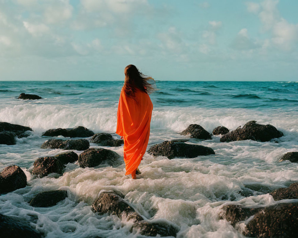 Orange-clothed person on rocky shoreline with crashing waves and cloudy sky