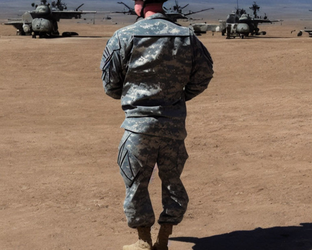 Soldier in camouflage uniform observing helicopters and military vehicles in desert setting