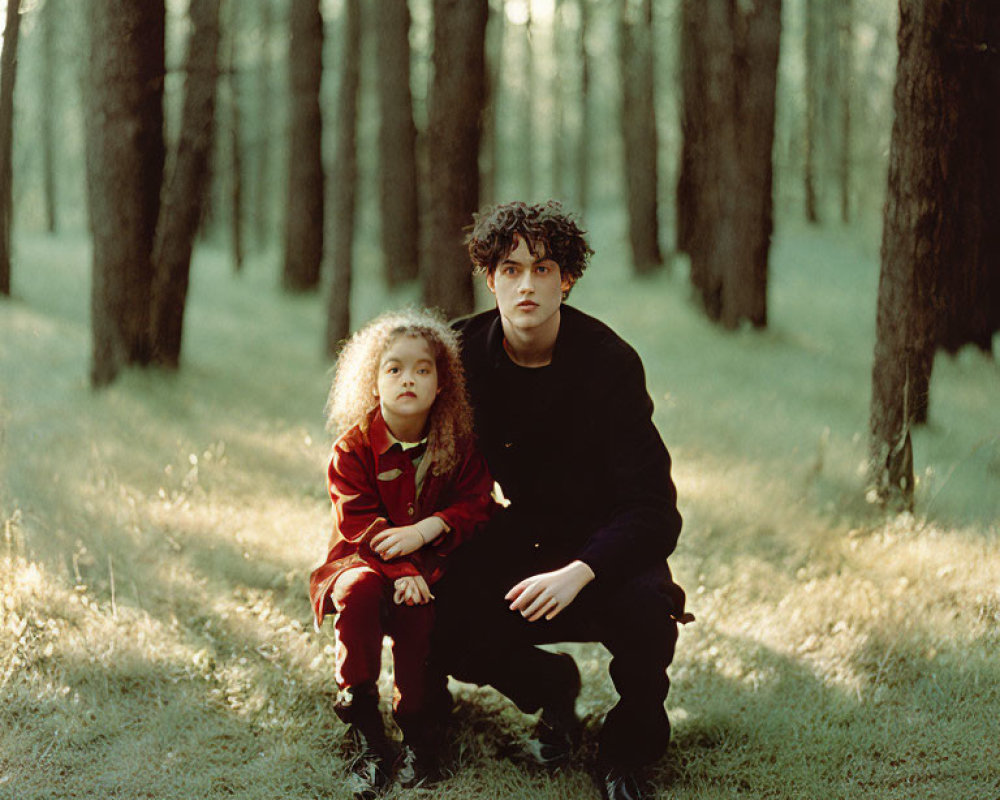 Adult and child squatting in sunlit forest: adult in black, child in red.