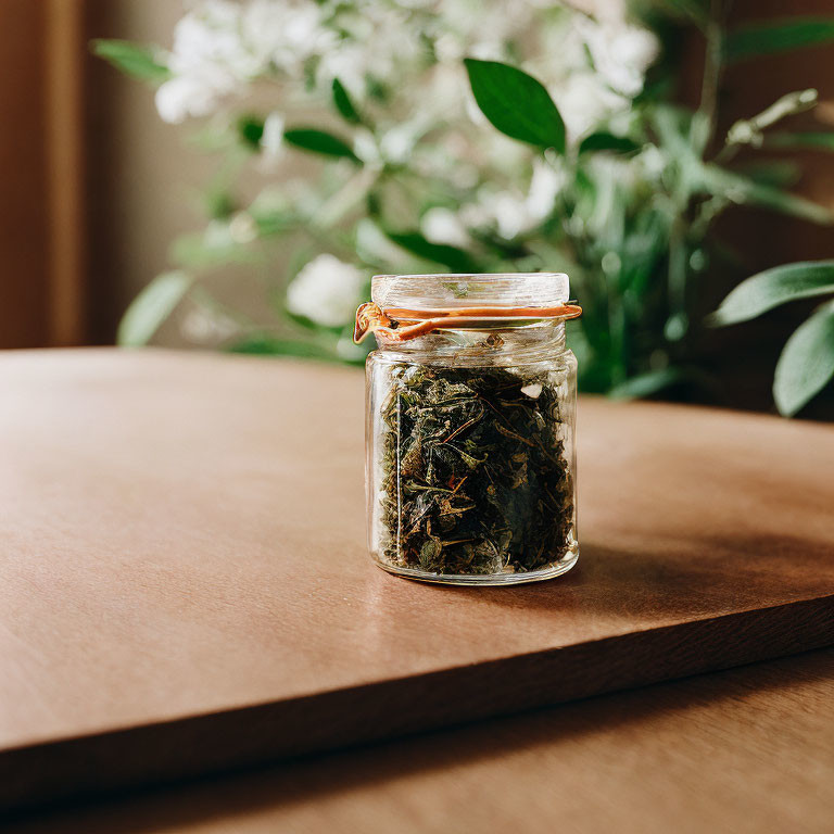 Glass jar filled with dried green tea leaves on wood table with blurred background of white flowers and greenery