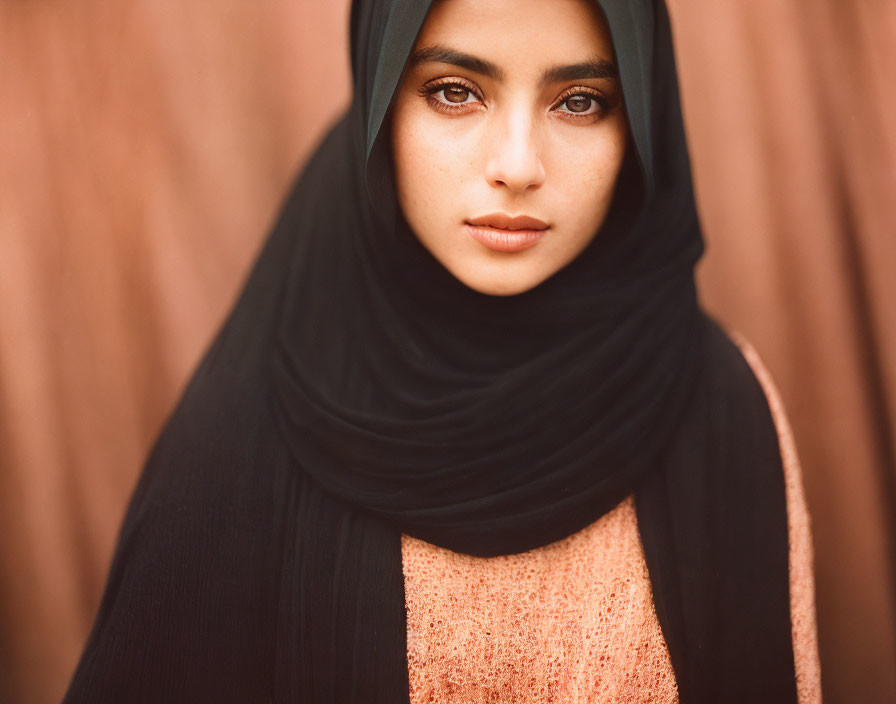 Woman portrait with black headscarf, intense gaze, coral top, blurry brown background