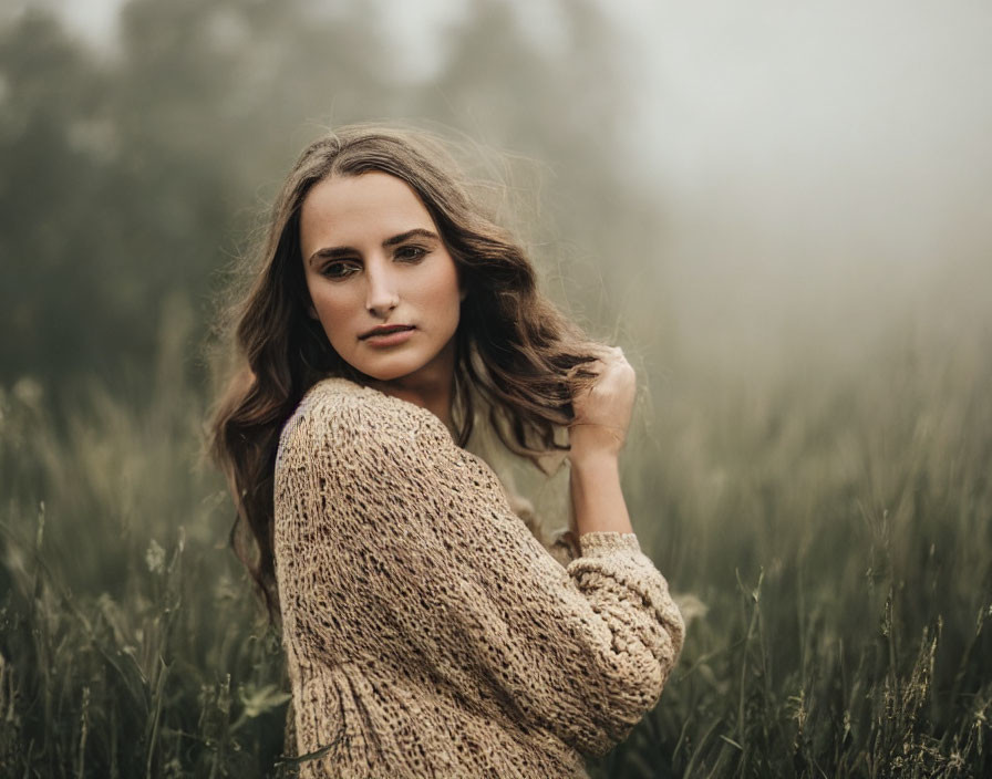 Woman in beige sweater standing in misty field gazes over shoulder.