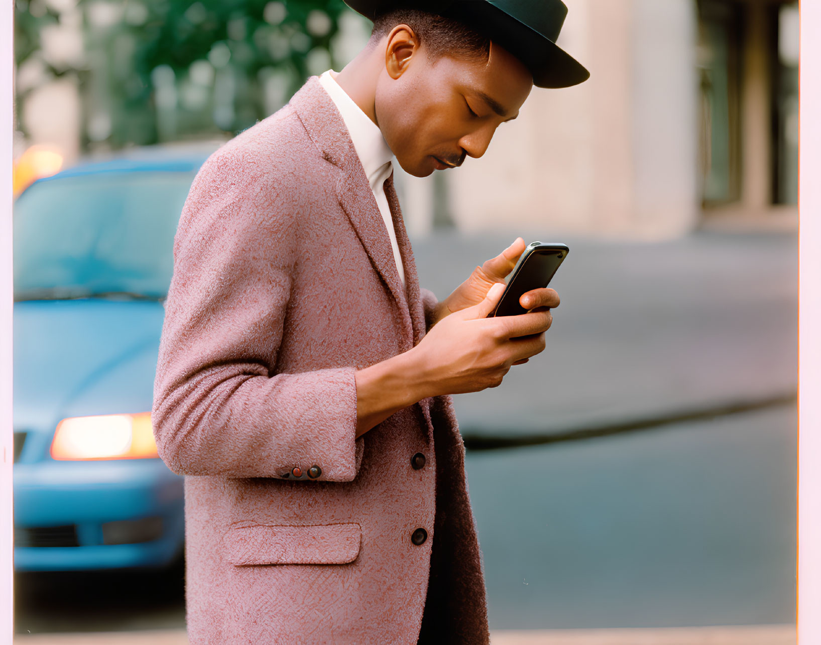 Fashionable man in pink coat and fedora hat on city street with smartphone