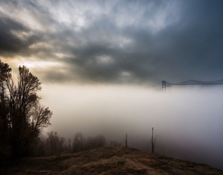 Foggy landscape at dawn or dusk with silhouettes of trees and bridge.