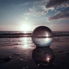 Surreal split reflective sphere on muddy terrain with smoky sky and sunset over water.