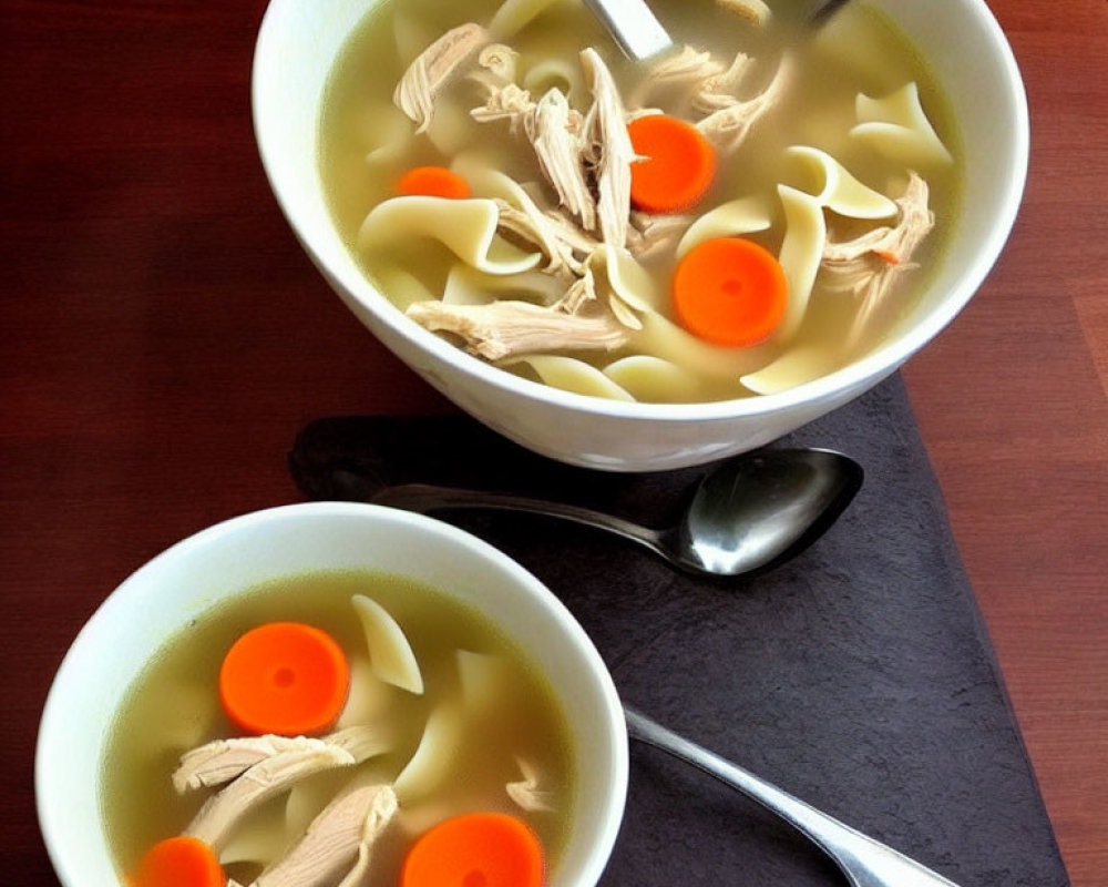 Chicken noodle soup bowls with carrots on dark table, each with spoon