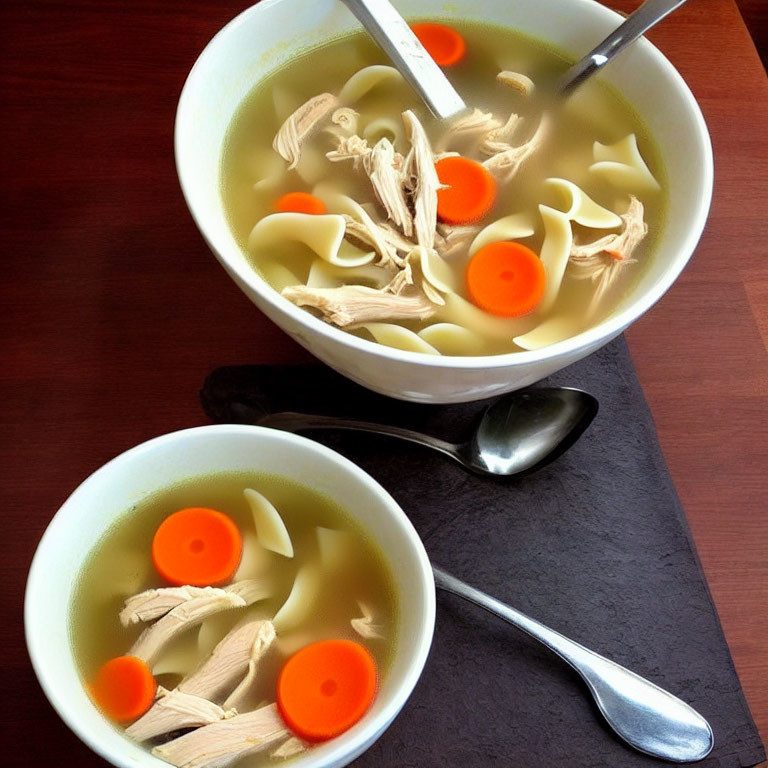 Chicken noodle soup bowls with carrots on dark table, each with spoon