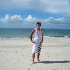 Man in white attire on sandy beach with ocean and sailboat in background