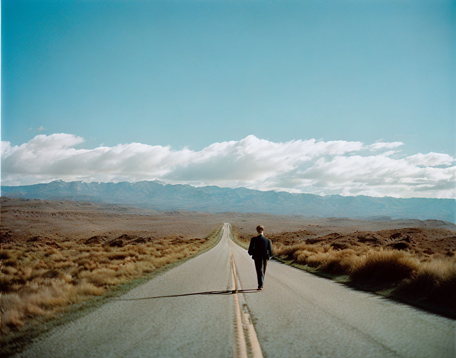 Lonely figure walking on straight road in barren landscape with distant mountains under cloudy sky