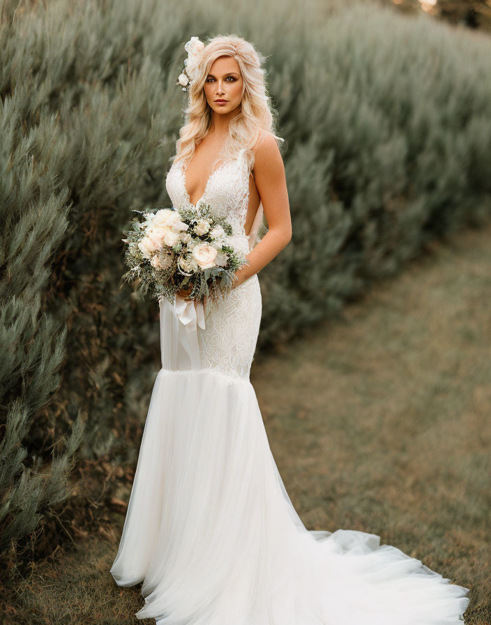 Bride in white lace gown with deep V-neckline holding bouquet against greenery backdrop