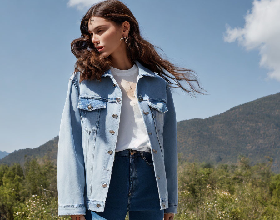 Woman in denim jacket outdoors with mountains and clear sky.