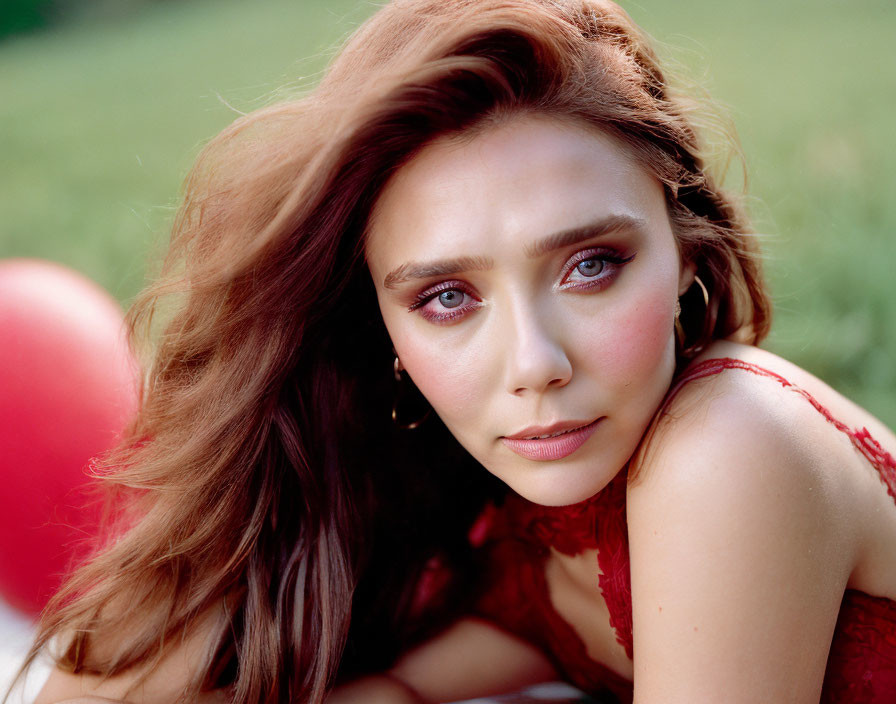 Woman with long brown hair and striking makeup posing on grass with red balloons in background.