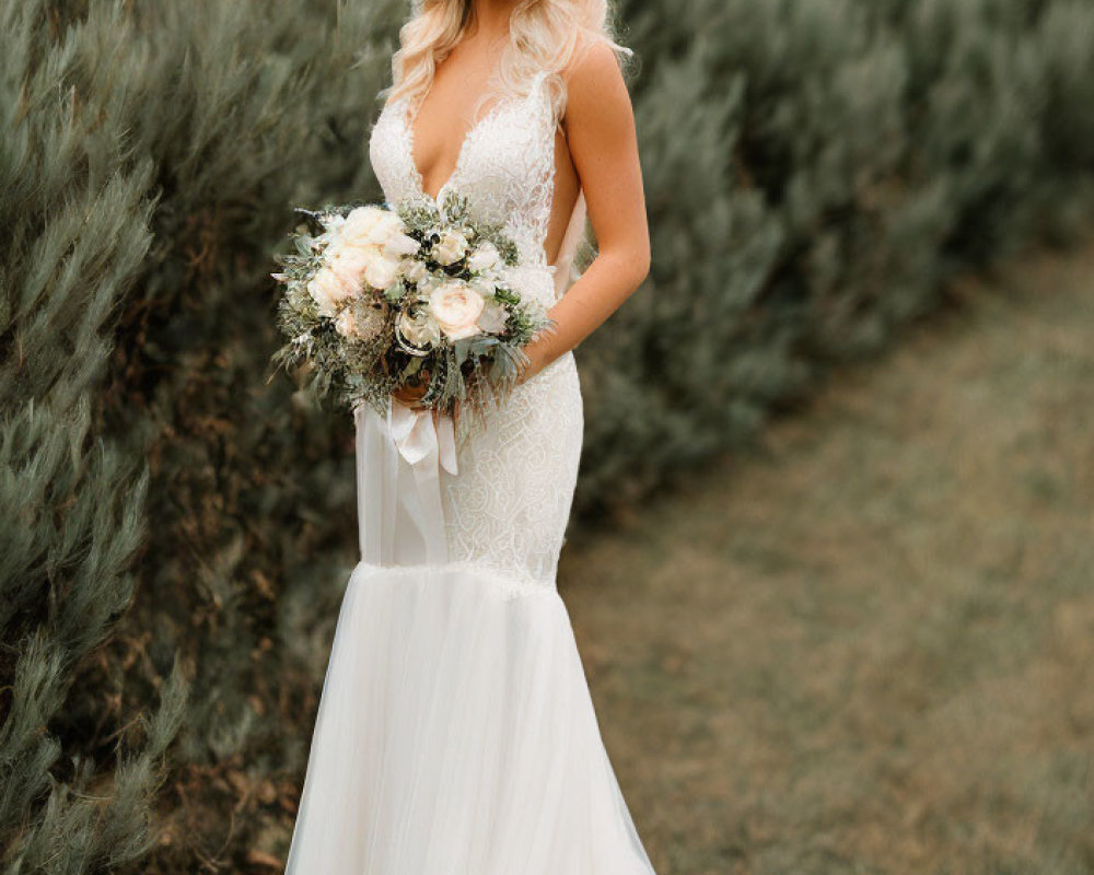 Bride in white lace gown with deep V-neckline holding bouquet against greenery backdrop