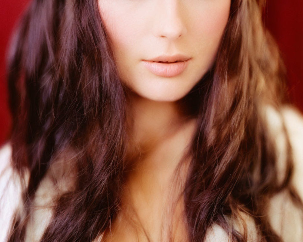 Woman with long wavy brown hair and blue eyes in white fur against red backdrop