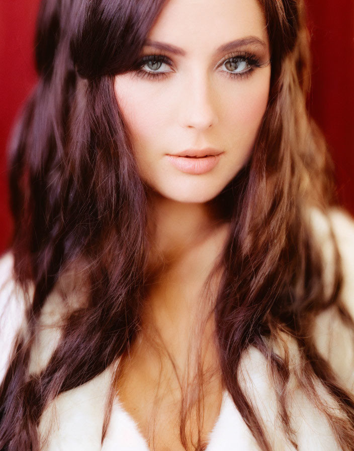 Woman with long wavy brown hair and blue eyes in white fur against red backdrop