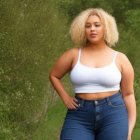 Blonde curly-haired woman in white tank top and blue jeans in front of lush greenery