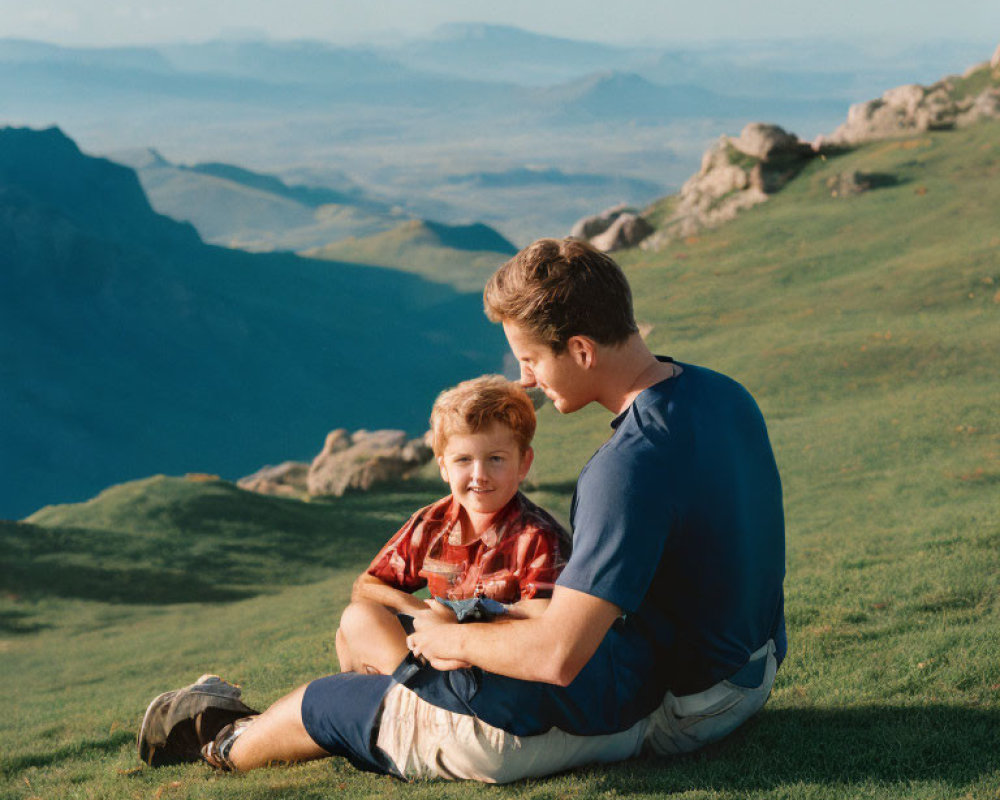 Man and boy on grassy hillside with vast mountain landscape.