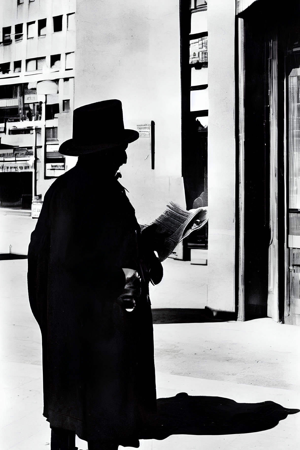 Figure in hat and coat reading newspaper by building column with sharp shadows