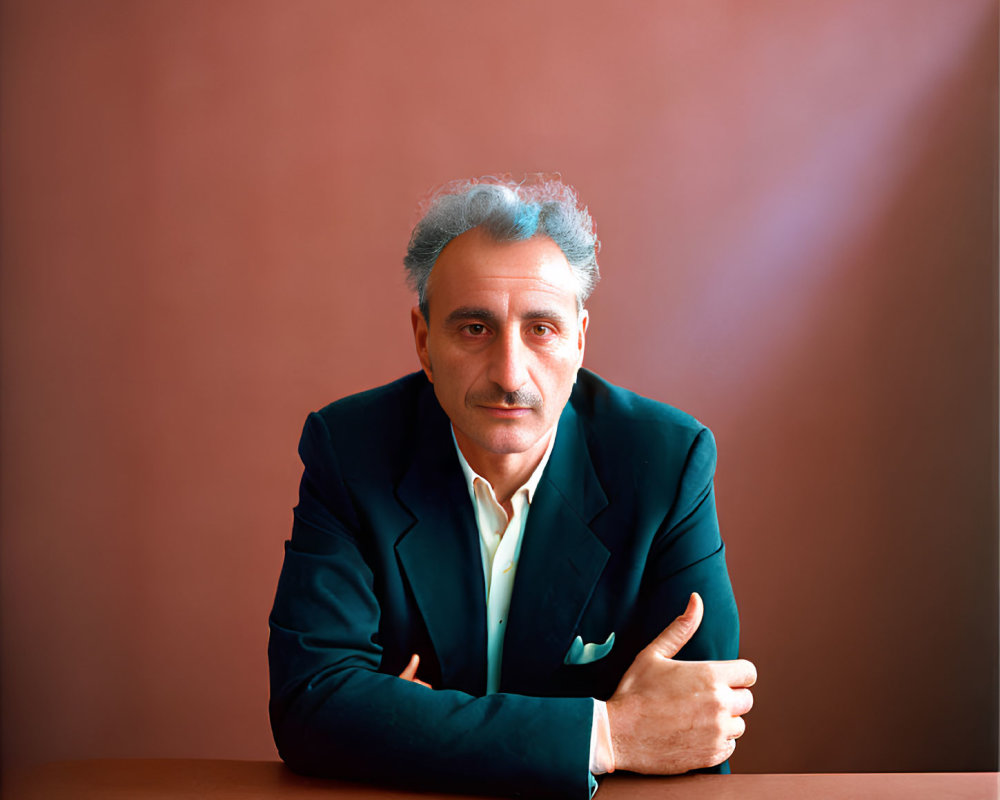 Salt-and-Pepper-Haired Man in Dark Suit and Green Shirt Seated Against Maroon Back