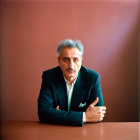 Salt-and-Pepper-Haired Man in Dark Suit and Green Shirt Seated Against Maroon Back