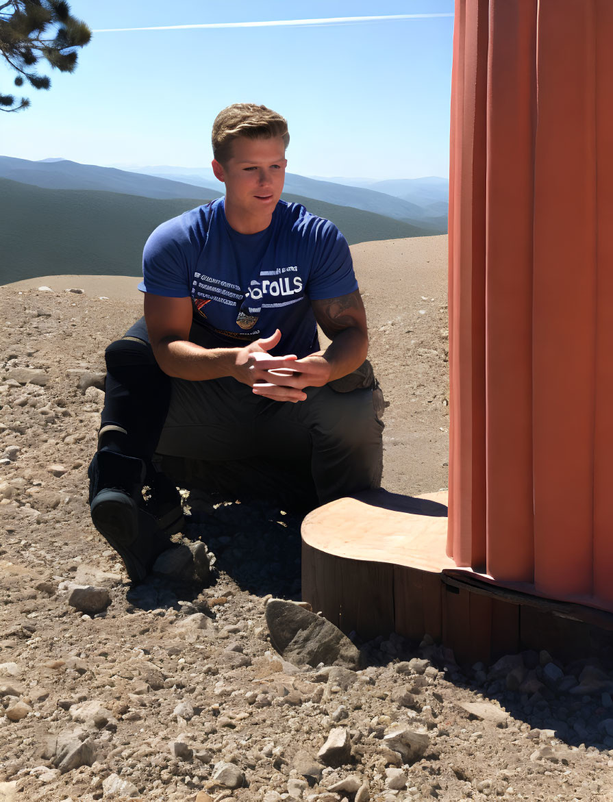 Man in Blue T-Shirt Sitting Outdoors with Phone and Mountain View