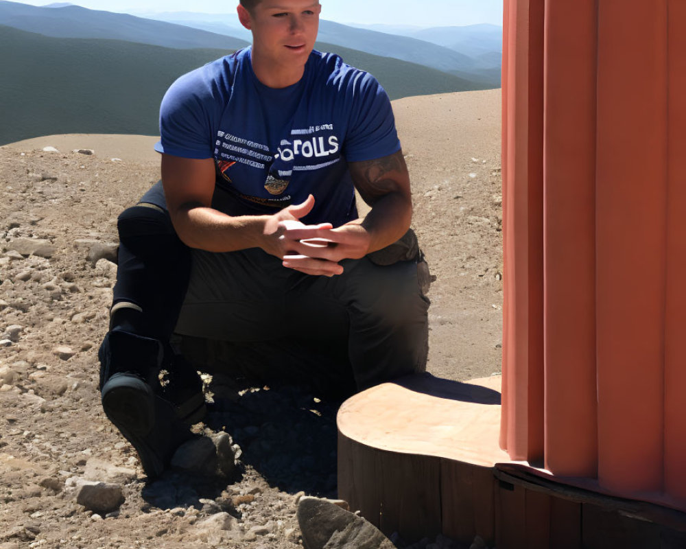 Man in Blue T-Shirt Sitting Outdoors with Phone and Mountain View