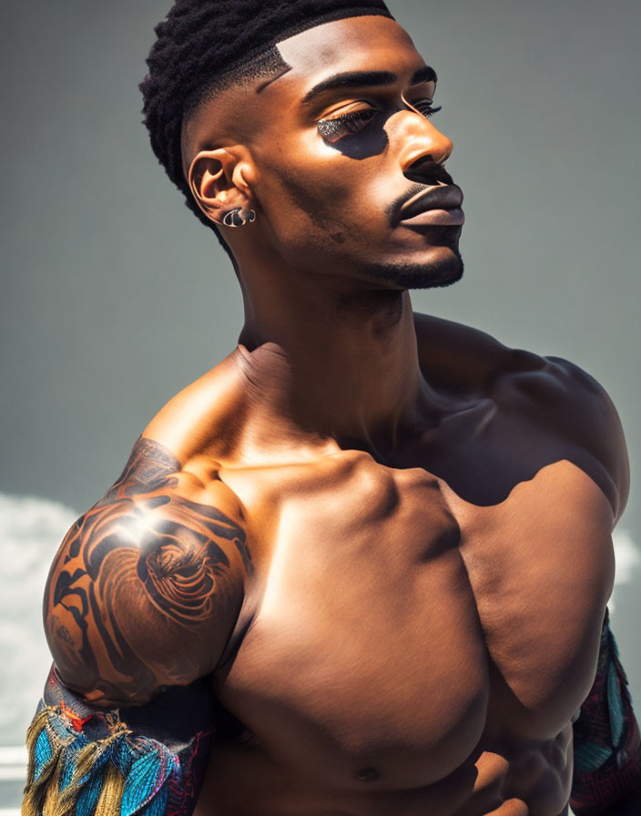 Muscular shirtless man with arm tattoo in pensive pose on soft backdrop