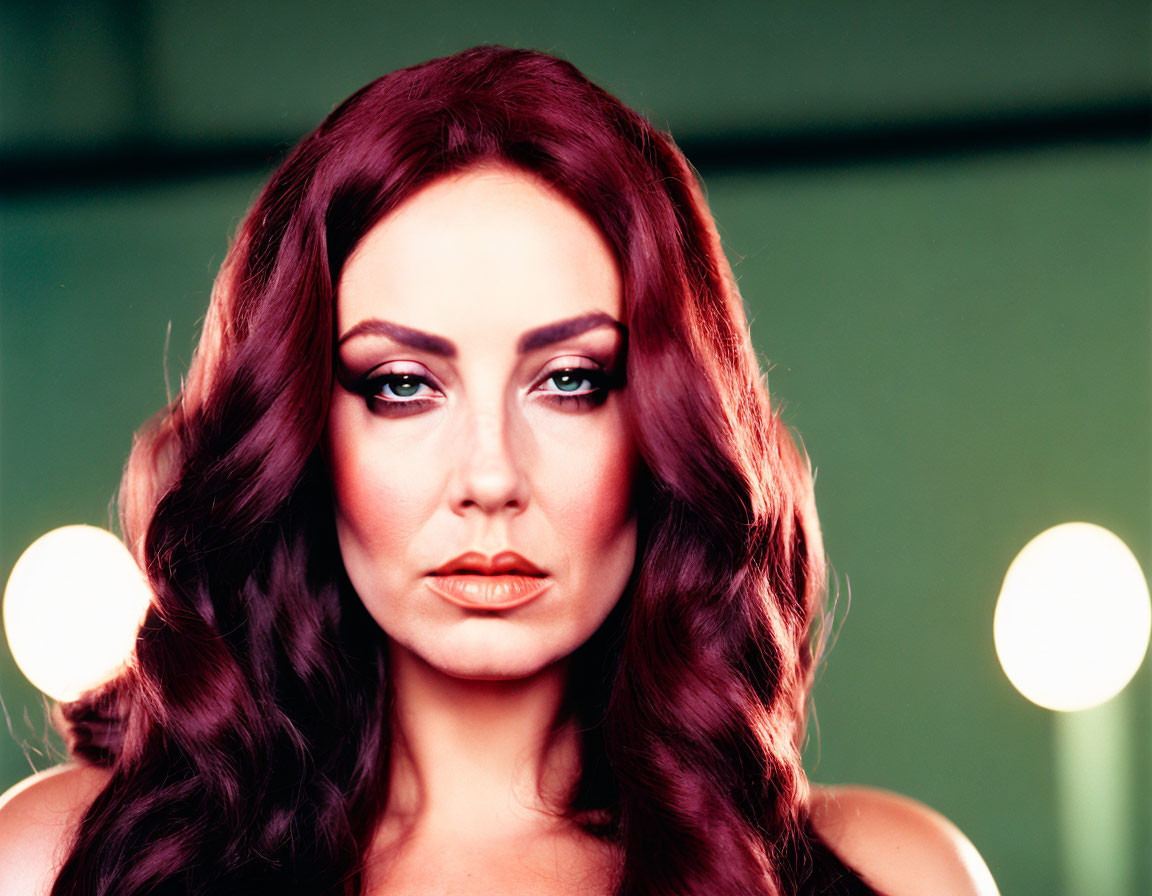 Portrait of woman with wavy auburn hair and makeup in front of blurred lights.