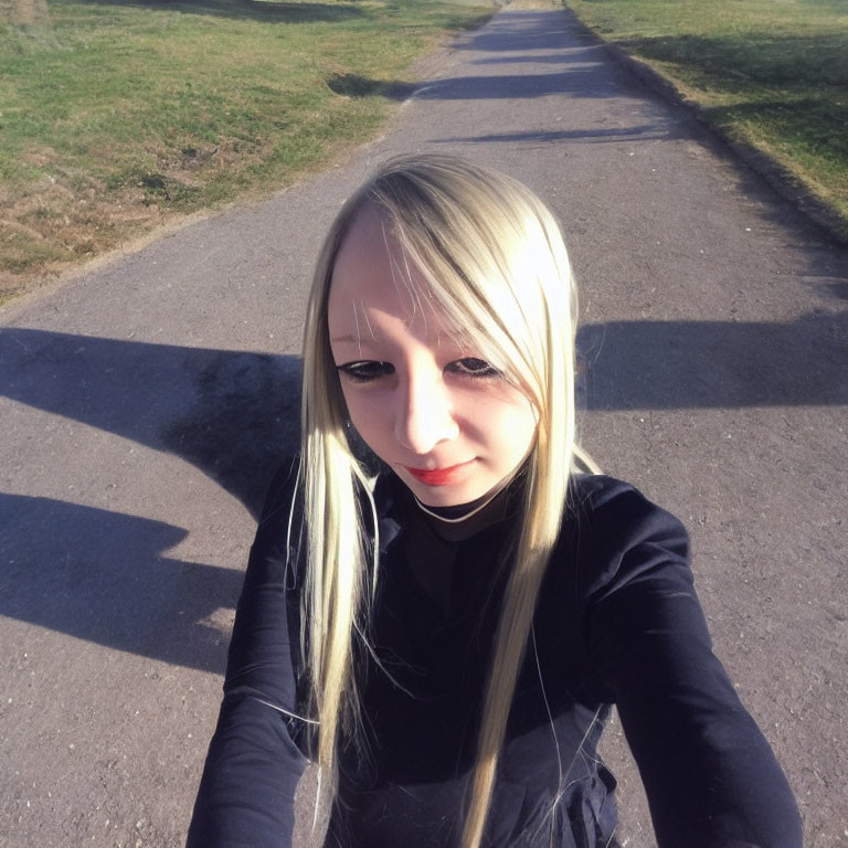 Blonde person in black top taking selfie on sunny grassy path