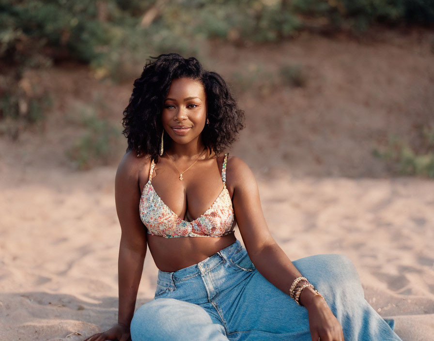 Curly-haired woman in patterned bikini top and denim pants on sand